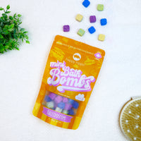 Bag of orange Kaleidoscope Mini Bath Bombs on a white background with mini bath bombs scattered around it