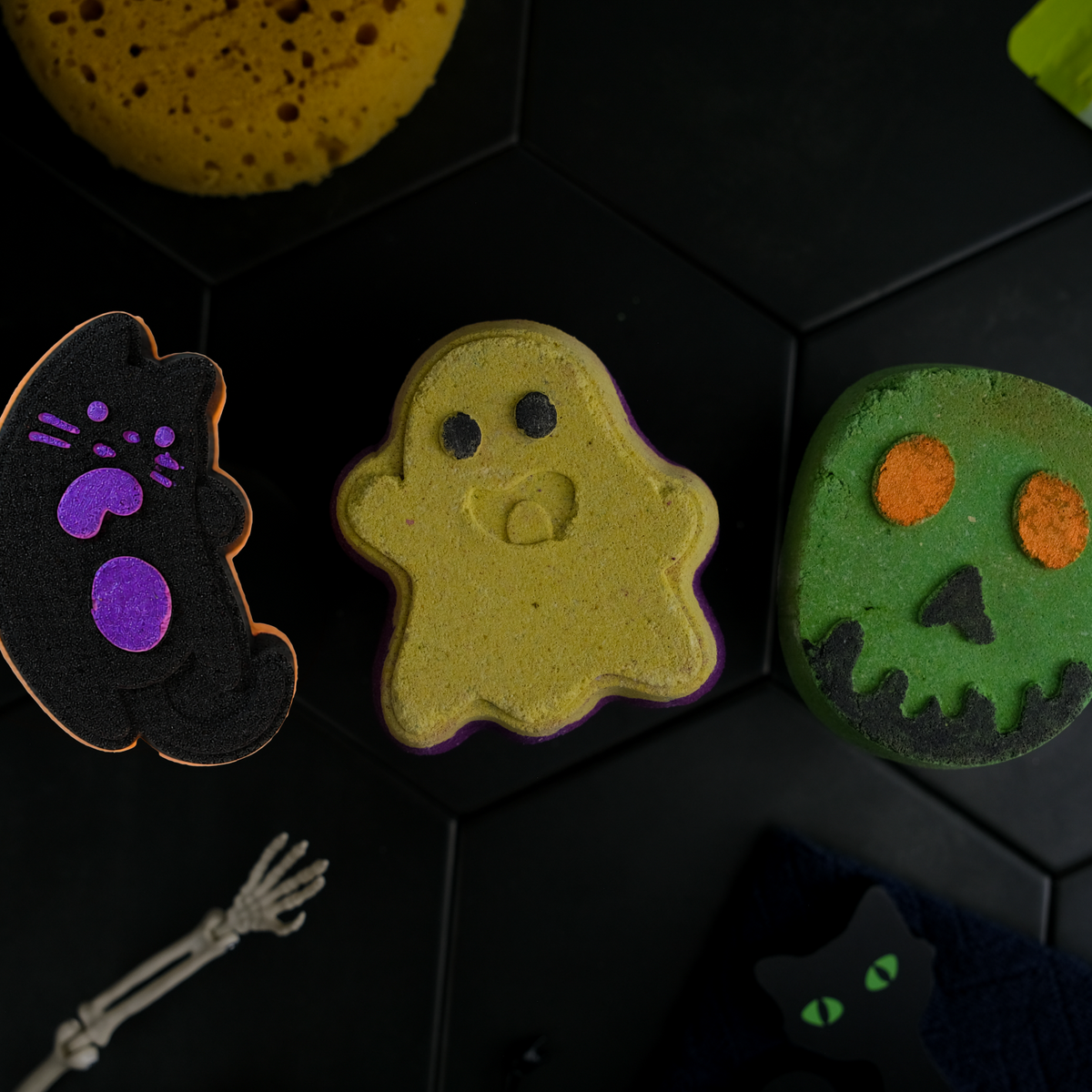 Three Halloween-themed bath bombs shaped like a black cat, a yellow ghost, and a green zombie, arranged on a dark hexagonal surface with other bath product packaging and props around them.
