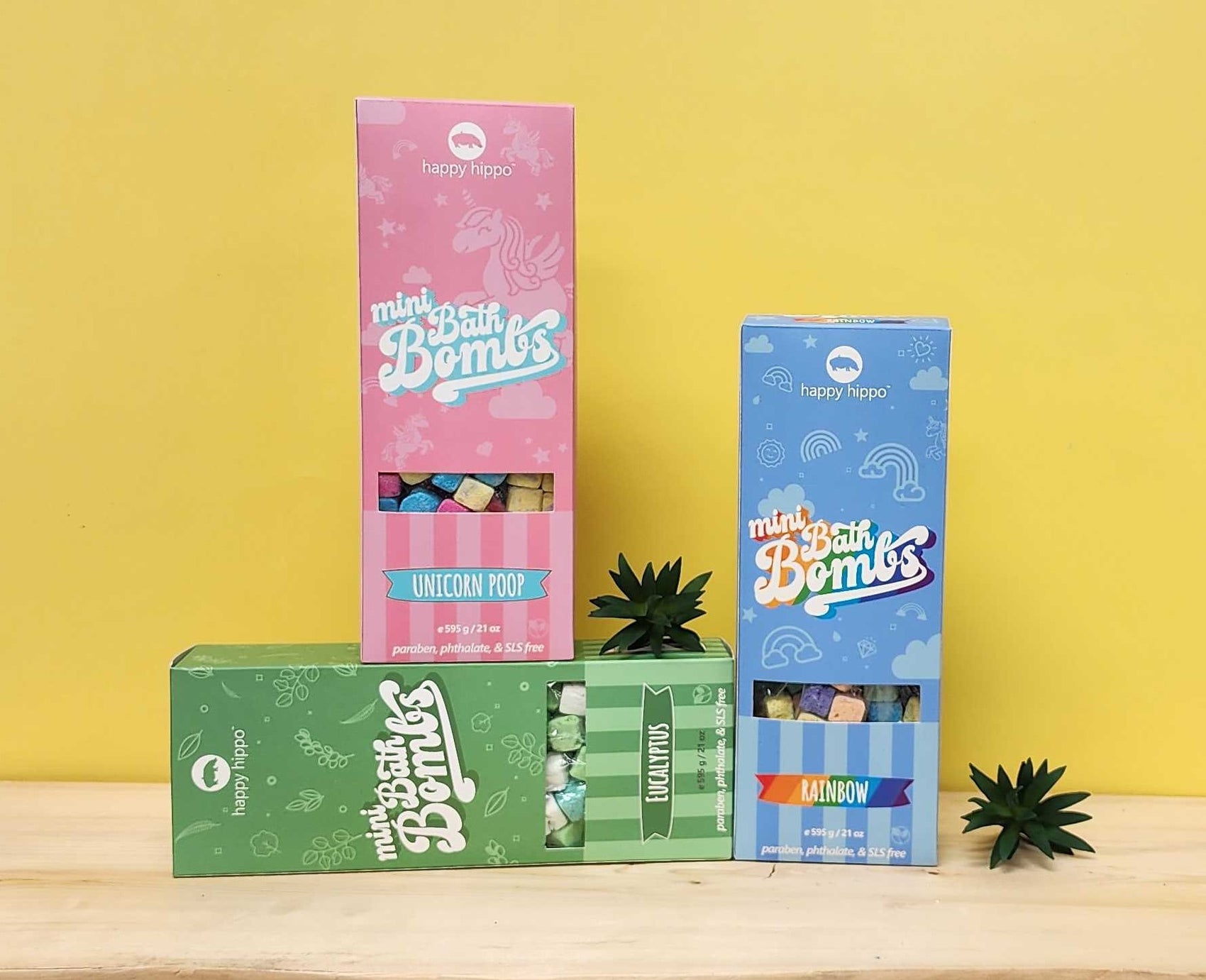 Three colorful boxes of 'Happy Hippo' bath bombs on a wooden surface with a yellow background.